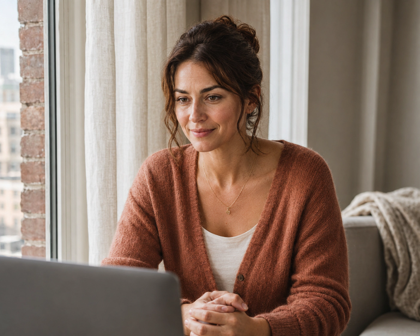 A woman in a rust cardigan smiling toward a laptop.