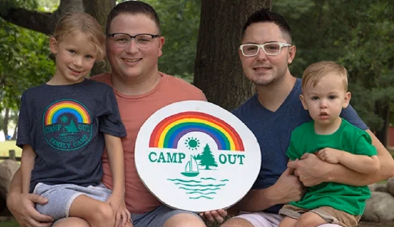 Two dads with their daughter and son smile holding a "Camp Out" sign.