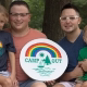 Two dads with their daughter and son smile holding a "Camp Out" sign.