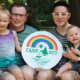 two dads hold their daughter and son with a "Camp Out" sign.