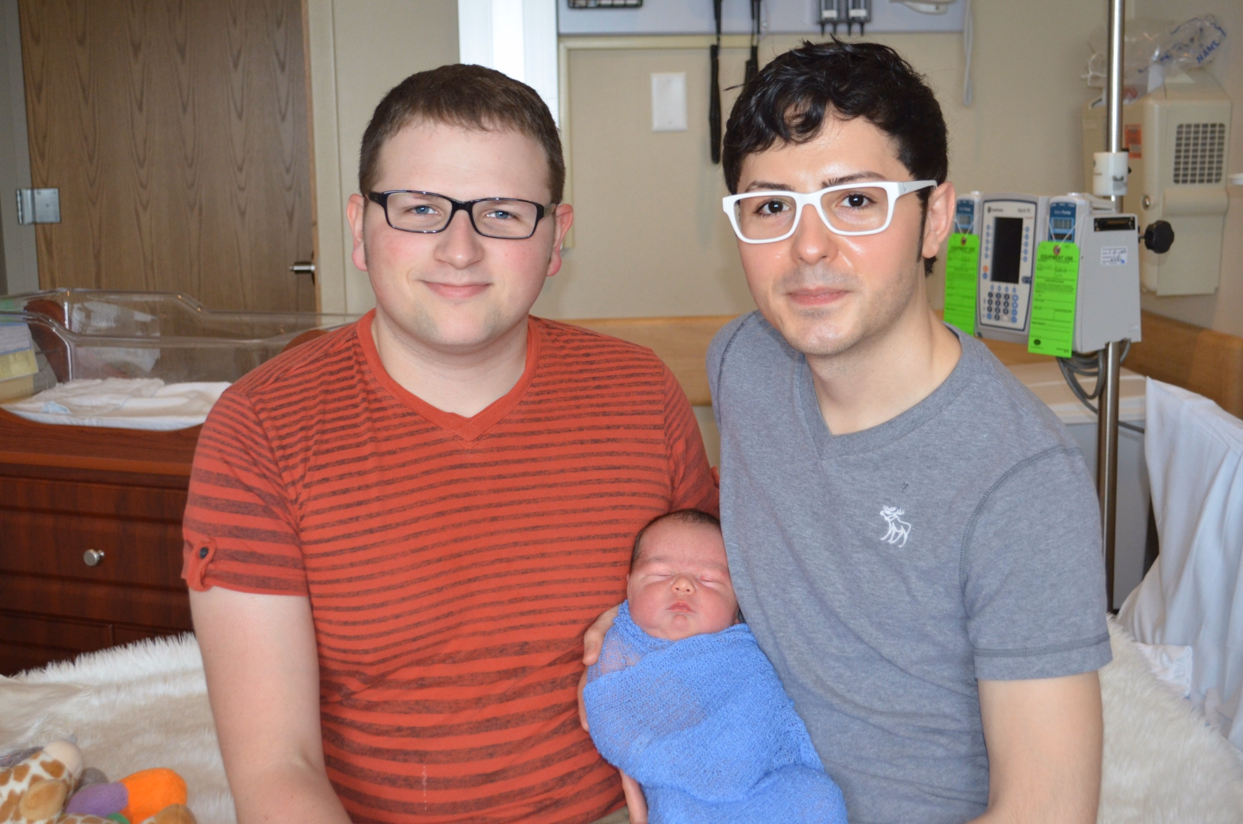 Two dads smile while they hold their son at a hospital.