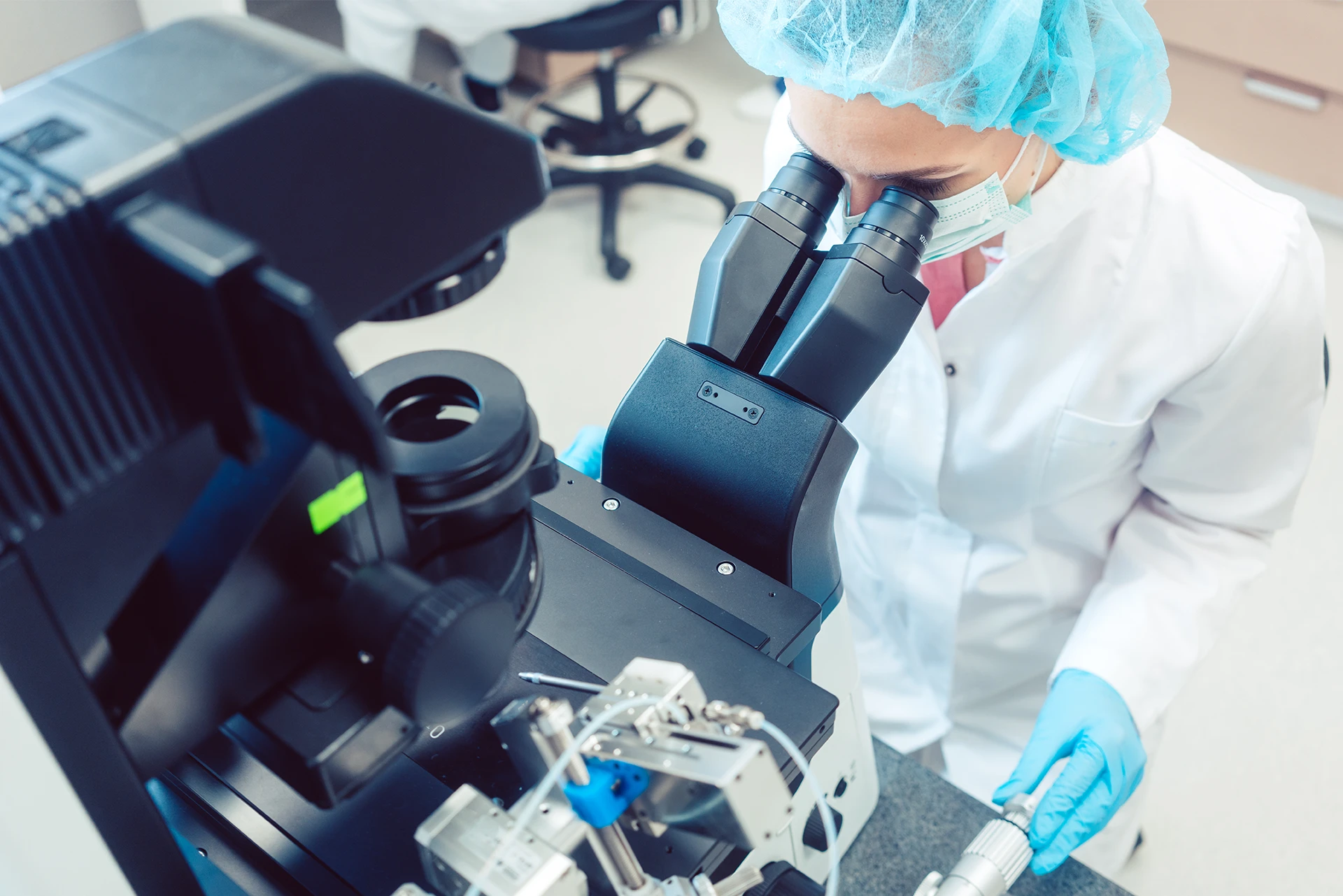 A woman in a white lab coat, blue gloves, and a blue hair net looking through a microscope.