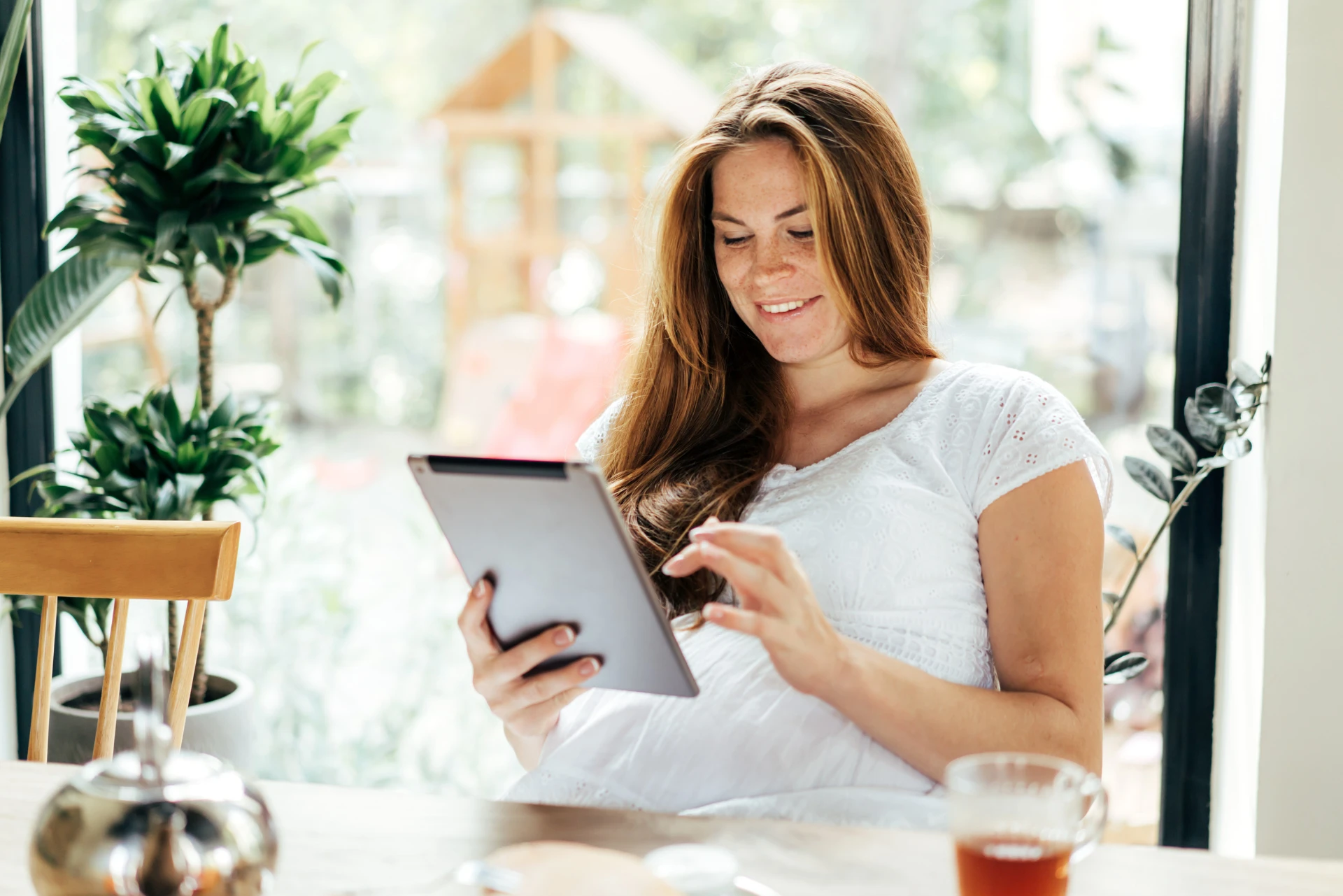 A pregnant woman in a white shirt sitting at a table, smiling while holding a tablet in her hand. 